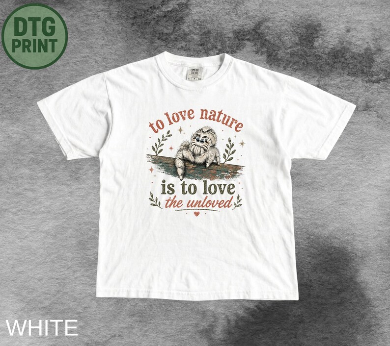 Może przedstawiać: Biały t-shirt z kr&oacute;tkim rękawem z grafiką skaczącego pająka na gałęzi. Napis "to love nature is to love the unloved" wykonany jest czerwoną i ciemnozieloną czcionką, ozdobiony małymi ilustracjami liści oraz małym czerwonym sercem.