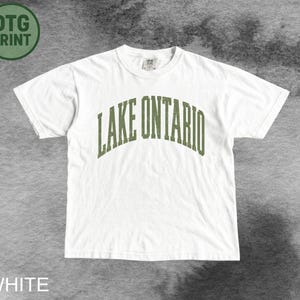 May include: A white short-sleeved t-shirt featuring the text LAKE ONTARIO printed in an arched, distressed olive green font across the chest. The garment has a crew neckline and a standard fit, with a small tag visible at the inner collar.