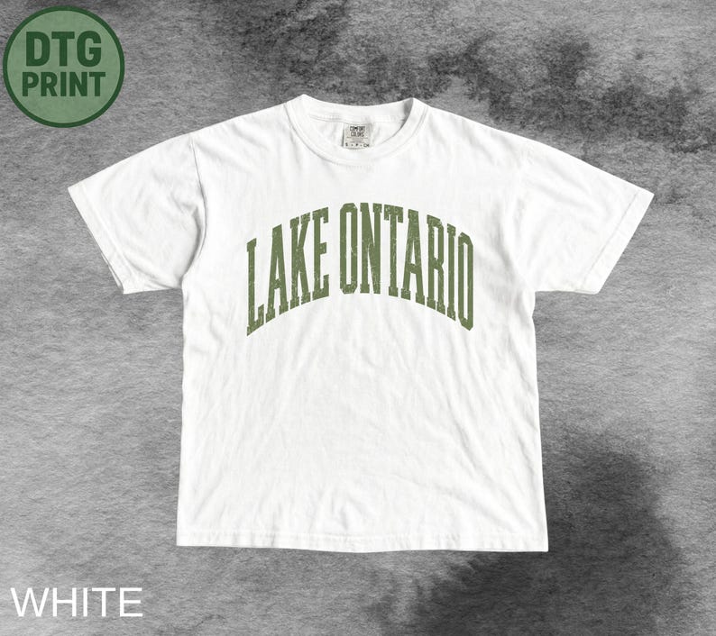 May include: A white short-sleeved t-shirt featuring the text LAKE ONTARIO printed in an arched, distressed olive green font across the chest. The garment has a crew neckline and a standard fit, with a small tag visible at the inner collar.