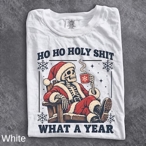 May include: A white t-shirt featuring a graphic of a skeleton dressed as Santa Claus sitting in a wooden chair, holding a red mug. The text reads HO HO HOLY SHIT WHAT A YEAR. The design includes snowflakes and stars in a dark blue ink color.