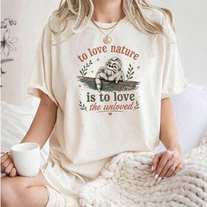 Może przedstawiać: Kremowy t-shirt z grafiką przedstawiającą małego pająka siedzącego na gałęzi. Napis głosi, to love nature is to love the unloved, a ilustrację otaczają drobne akcenty w kształcie gwiazdek i liści w odcieniach brązu oraz zieleni.