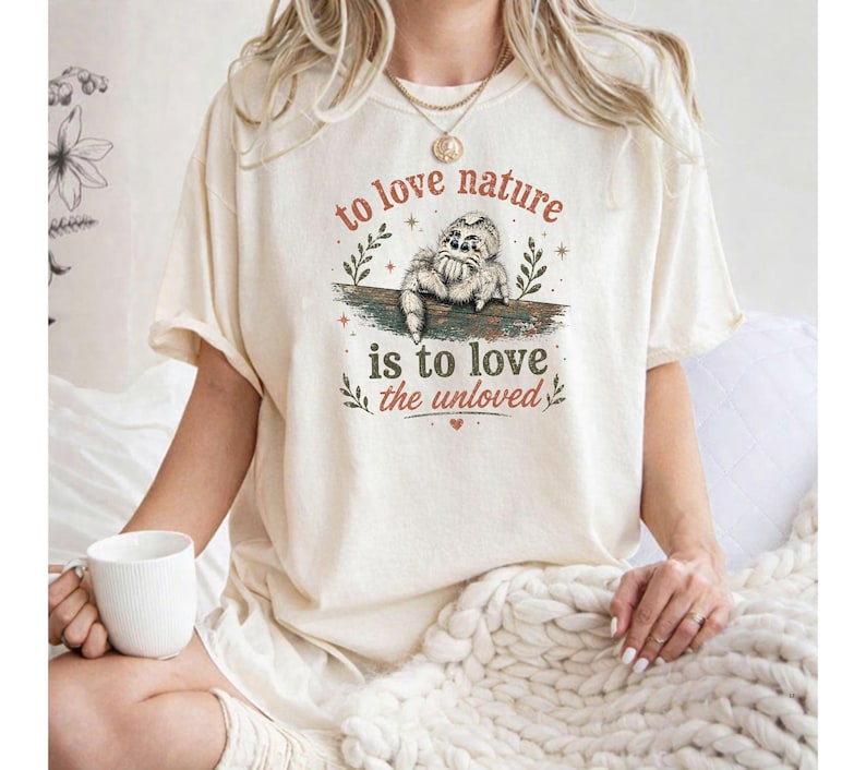 Może przedstawiać: Kremowy t-shirt z grafiką przedstawiającą małego pająka siedzącego na gałęzi. Napis głosi, to love nature is to love the unloved, a ilustrację otaczają drobne akcenty w kształcie gwiazdek i liści w odcieniach brązu oraz zieleni.