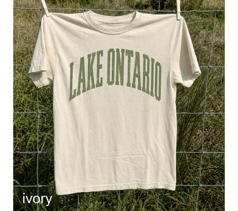 May include: An ivory short-sleeve t-shirt featuring the text LAKE ONTARIO printed in a distressed, arched green font across the chest. The shirt has a crew neckline and a standard fit, showcasing a clean design with a vintage-inspired graphic.