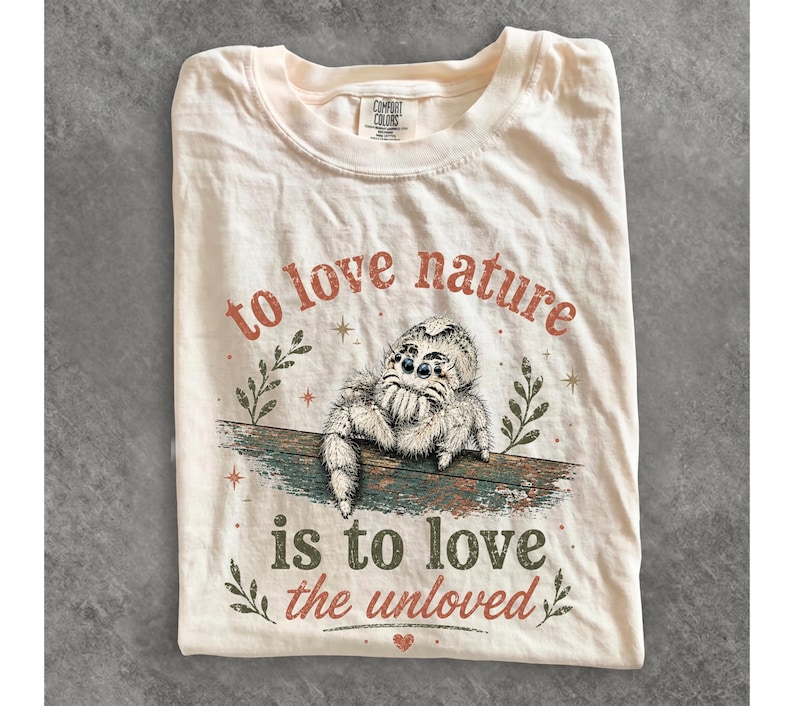 Może przedstawiać: Kremowy t-shirt z grafiką puszystego pająka z rodziny skakunowatych siedzącego na gałęzi. Napis głosi "to love nature is to love the unloved", a całość uzupełniają małe ilustracje liści i gwiazd w stonowanych odcieniach czerwieni i zieleni.