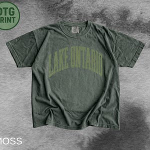 May include: A moss green short-sleeve t-shirt featuring the text LAKE ONTARIO in a light green arched font across the chest. The garment has a crew neckline and a relaxed fit, with a small tag visible at the inner collar area of the shirt.