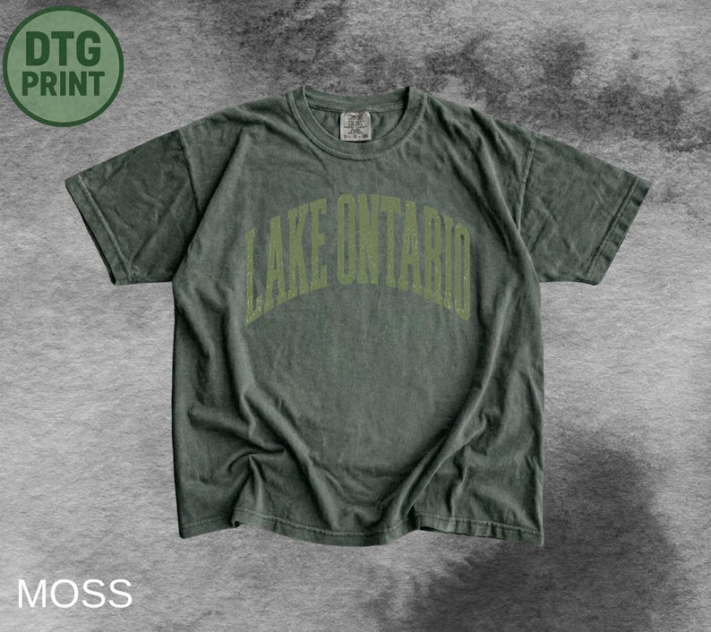 May include: A moss green short-sleeve t-shirt featuring the text LAKE ONTARIO in a light green arched font across the chest. The garment has a crew neckline and a relaxed fit, with a small tag visible at the inner collar area of the shirt.