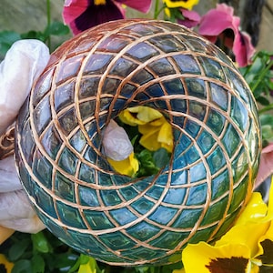 Könnte beinhalten: Eine teal- und kupferfarbene, geometrische Harz-Kugel mit einem runden Loch in der Mitte. Die Kugel ist von lila, gelben und orangefarbenen Blumen umgeben.