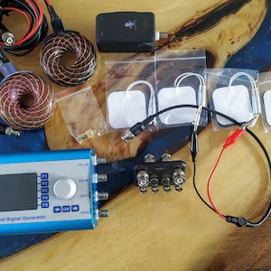 Può includere: Un generatore di segnale a 2 canali blu e argento con uno schermo che mostra "Spooky2" e una grafica di un fantasma. Il dispositivo ha una manopola, pulsanti e più porte. L'immagine include anche due bobine, una scatola nera e quattro elettrodi bianchi.