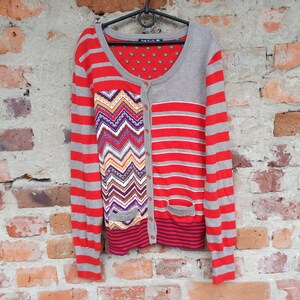 May include: A grey and red striped cardigan with a colourful chevron pattern on the front. The cardigan has a button closure and a pocket on the left side.