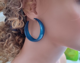 May include: A pair of large, glossy, teal-colored hoop earrings. The earrings are a bold, crescent shape, with a smooth, reflective surface. The hoops are worn on a person's ear, adding a pop of color.