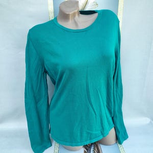 Puede incluir: Un suéter de manga larga de color verde azulado con cuello redondo. El suéter se exhibe en un maniquí. La tela parece ser un material de punto. Las mangas son largas y el dobladillo es recto.
