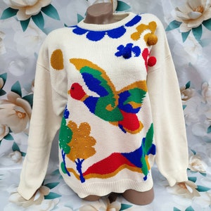May include: A cream-coloured long-sleeve knit jumper featuring a colourful intarsia design of a flying duck, trees, and hills. The jumper is accented with blue scalloped trim at the neckline and three-dimensional pom-poms in gold, blue, and red.