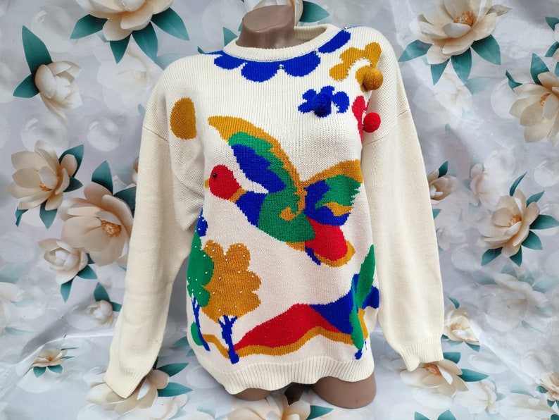 May include: A cream-coloured long-sleeve knit jumper featuring a colourful intarsia design of a flying duck, trees, and hills. The jumper is accented with blue scalloped trim at the neckline and three-dimensional pom-poms in gold, blue, and red.