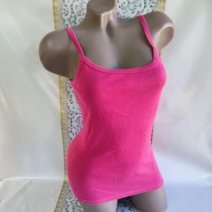 May include: A bright pink camisole top with thin straps and a scoop neckline. The fitted tank top is displayed on a mannequin. The fabric appears to be a soft, stretchy material. The background is a neutral white and gold.