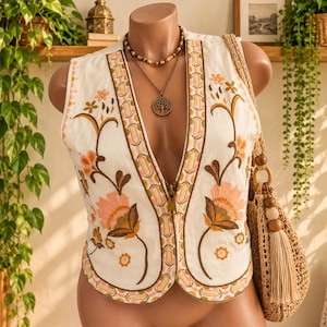 May include: An off-white sleeveless waistcoat featuring intricate floral embroidery in shades of peach, brown, and gold. The garment has a deep V-neckline with a central zip closure and a decorative border pattern along the edges and hemline.