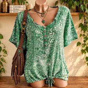 May include: A green short-sleeved blouse featuring a white abstract floral pattern, a button-down placket, and a drawstring hem. The blouse is paired with a brown leather shoulder bag with fringe detailing and a layered beaded necklace.