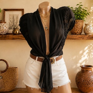 May include: A black sheer blouse with short puffed sleeves and a front tie closure, paired with white denim shorts featuring a raw hem and a brown leather belt with a gold-toned buckle. The blouse has a deep V-neckline and ruched sleeve details.