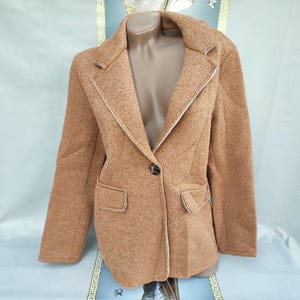 Puede incluir: Un blazer marrón claro de tejido de punto texturizado. El blazer tiene solapas con muescas, dos bolsillos delanteros y cierre de un solo botón. Ribetes blancos acentúan las solapas y los bolsillos. El blazer se muestra en un maniquí.