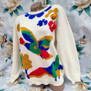 May include: A cream-coloured long-sleeve knit jumper featuring a colourful intarsia design of a flying duck, trees, and hills in blue, green, red, and yellow. The jumper includes three-dimensional pom-poms and a ribbed crew neckline.