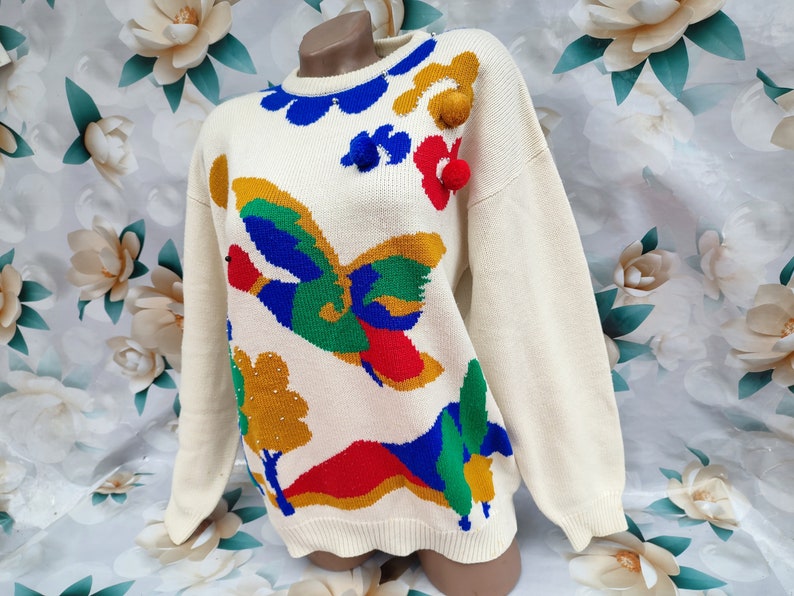 May include: A cream-coloured long-sleeve knit jumper featuring a colourful intarsia design of a flying duck, trees, and hills in blue, green, red, and yellow. The jumper includes three-dimensional pom-poms and a ribbed crew neckline.