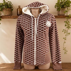 May include: A brown zip-up hoodie featuring a repeating white star pattern, a white sherpa-lined hood with white drawstrings, long sleeves with brown cuffs, and a circular patch on the upper left chest. The garment has a solid brown lower panel.