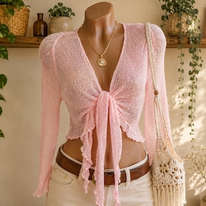 May include: A light pink long-sleeve cardigan with a sheer, open-knit texture and a front tie closure. The garment features ruffled edges along the hem and tie ends. It is worn with a gold-toned pendant necklace, a brown leather belt, and white pants.