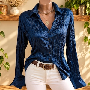 May include: A long-sleeved button-down shirt in a deep, metallic blue with a crinkled, textured finish. The shirt features a classic collar, a front button placket, and wide cuffs. It is paired with white trousers and a brown leather belt.