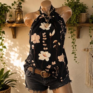 May include: A black halter-neck top featuring a white and light pink floral print, paired with dark grey denim shorts and a brown woven leather belt. The top has a high neckline and is worn with a gold-toned chain necklace with a circular pendant.