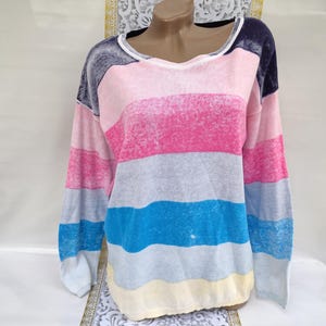 Może przedstawiać: Sweter z długim rękawem w poziome paski w odcieniach różu, szarości, błękitu i żółci. Sweter ma dekolt w serek i luźny krój. Rękawy są długie, a materiał wydaje się miękki i lekki. Kolorystyka jest jasna i wesoła.