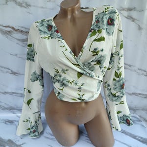 Può includere: Top a portafoglio color crema con maniche lunghe e scollo a V. Il top è adornato da un motivo floreale nei toni del blu e del verde, con accenti rossi. Le maniche hanno un design svasato, che aggiunge un tocco di eleganza all'indumento.