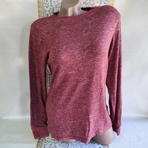 May include: A long-sleeved maroon shirt with a crew neck. The fabric has a heathered texture, with lighter flecks throughout the maroon material. The sleeves end in a ribbed cuff. The shirt is displayed on a mannequin.