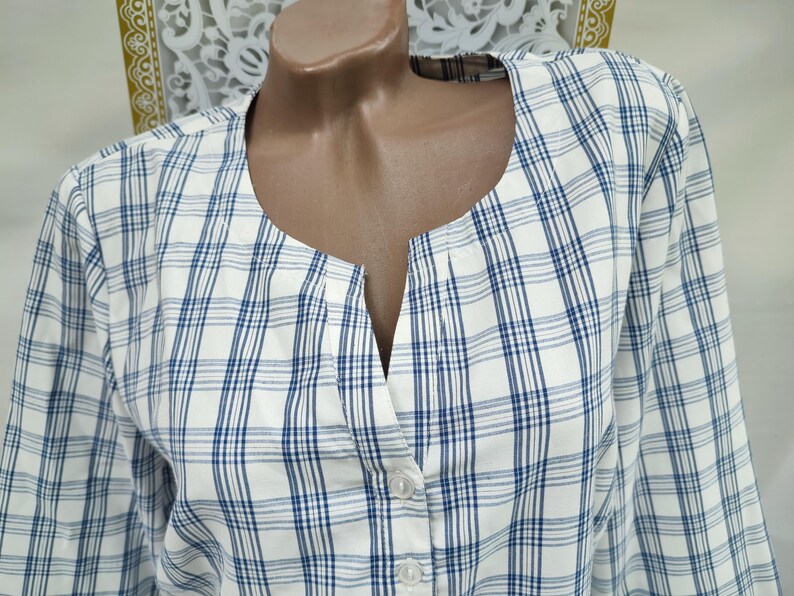 Pu&ograve; includere: Camicia a maniche lunghe a quadri bianchi e blu con scollatura a V e chiusura con bottoni. La camicia &egrave; esposta su un manichino. Il motivo a quadri presenta sottili linee blu su sfondo bianco.