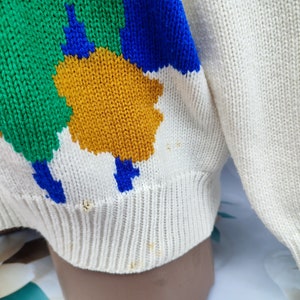 May include: A cream-coloured knit jumper featuring a bold, abstract pattern of large, irregular shapes in vibrant green, royal blue, and mustard yellow. The garment has a ribbed hem and cuffs, with visible small stains and minor fraying near the bottom.