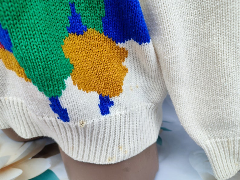May include: A cream-coloured knit jumper featuring a bold, abstract pattern of large, irregular shapes in vibrant green, royal blue, and mustard yellow. The garment has a ribbed hem and cuffs, with visible small stains and minor fraying near the bottom.