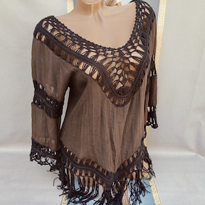 Puede incluir: Top de ganchillo marrón con flecos. El top presenta un escote amplio y abierto y detalles de ganchillo alrededor del escote y las mangas. Las mangas son largas y acampanadas, y el dobladillo está rematado con flecos.