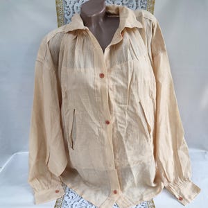 May include: A light peach-colored button-down shirt with long sleeves. The shirt has a collared neckline, a front pocket on each side, and buttons down the front and on the cuffs. The fabric has a subtle textured pattern.