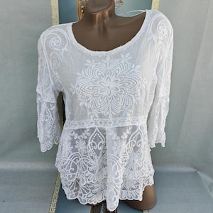 Puede incluir: Blusa blanca bordada con motivos florales. Cuello redondo y mangas tres cuartos. El bordado se concentra en el corpiño y las mangas, creando un aspecto delicado y calado. La tela es ligera y semitransparente, ideal para climas cálidos.