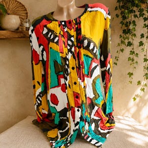 May include: A long-sleeved button-down shirt featuring a vibrant, abstract multi-coloured print. The design incorporates bold blocks of yellow, red, teal, black, and white in a geometric pattern. The shirt has a round neckline and vertical pleats.