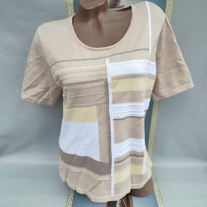 May include: Beige short-sleeve shirt with a scoop neck and a geometric design. The design features rectangular and striped patterns in shades of beige, white, and yellow. The shirt is displayed on a mannequin.