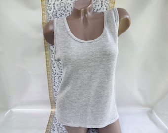 May include: A light gray tank top with a scoop neck and a textured knit pattern. The sleeveless top is displayed on a mannequin. The background includes a patterned fabric and a white surface.
