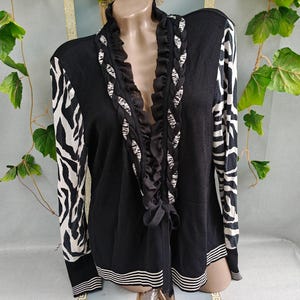 May include: A black cardigan featuring long sleeves with a white and black zebra print pattern. The front opening is adorned with a ruffled black trim and a braided detail in a zebra print. The hem and cuffs are finished with white horizontal stripes.