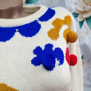 May include: A cream-coloured knit jumper featuring a crew neckline and a pattern of abstract shapes in royal blue and mustard yellow. The garment is embellished with small silver beads and three-dimensional pom-poms in mustard, blue, and red.