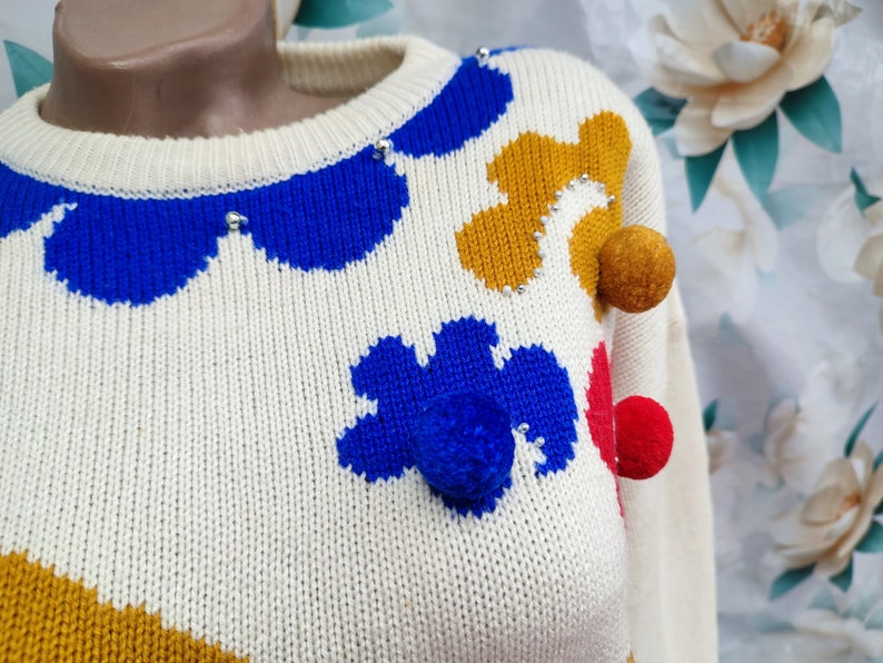 May include: A cream-coloured knit jumper featuring a crew neckline and a pattern of abstract shapes in royal blue and mustard yellow. The garment is embellished with small silver beads and three-dimensional pom-poms in mustard, blue, and red.