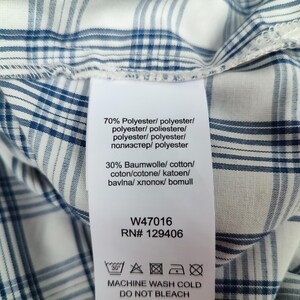 Pu&ograve; includere: Primo piano di un tessuto a quadri bianco e blu con un'etichetta. L'etichetta include i dettagli della composizione del tessuto: 70% poliestere e 30% cotone. L'etichetta include anche le istruzioni di lavaggio e il testo "MACHINE WASH COLD DO NOT BLEACH".