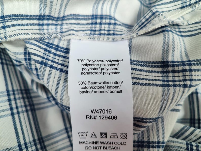 Pu&ograve; includere: Primo piano di un tessuto a quadri bianco e blu con un'etichetta. L'etichetta include i dettagli della composizione del tessuto: 70% poliestere e 30% cotone. L'etichetta include anche le istruzioni di lavaggio e il testo "MACHINE WASH COLD DO NOT BLEACH".