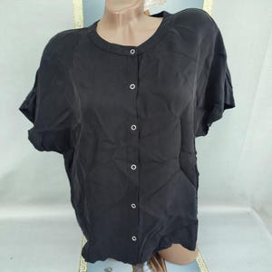 Puede incluir: Camisa negra de manga corta con cuello redondo y seis botones blancos. La tela parece ligera y ligeramente texturizada. La camisa se muestra en un maniquí, destacando su diseño sencillo y elegante.