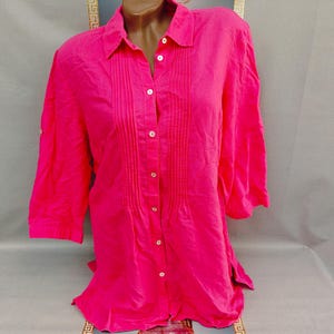 Puede incluir: Una camisa rosa brillante con botones, cuello y mangas tres cuartos. La camisa presenta pliegues verticales en la parte delantera y botones blancos. La tela parece ligera y adecuada para climas cálidos.