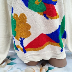 May include: A cream-coloured long-sleeve jumper featuring a knit landscape pattern with bold shapes in red, blue, green, and mustard yellow. The design includes stylised trees and hills, with small silver-toned beads accenting the mustard tree.