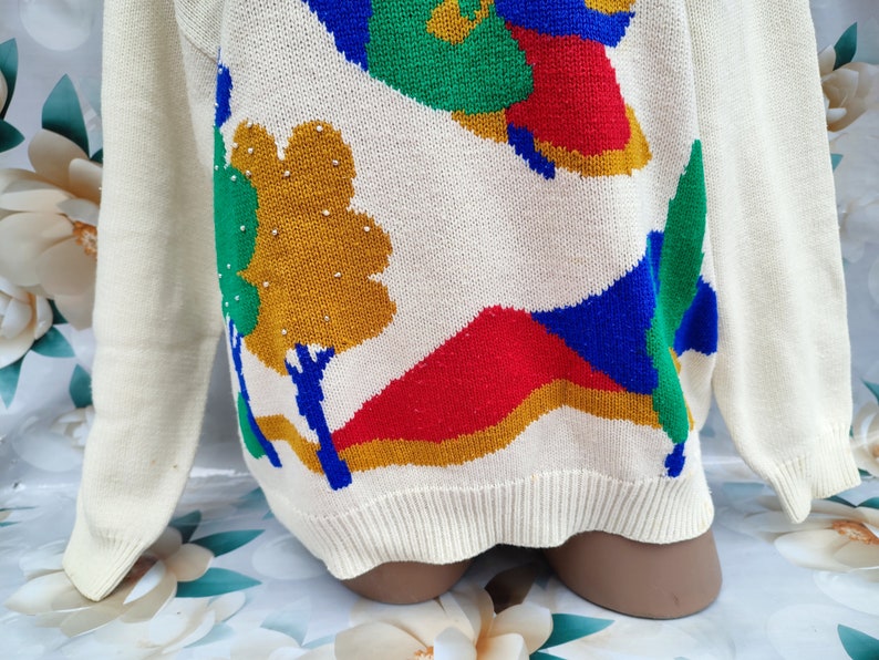 May include: A cream-coloured long-sleeve jumper featuring a knit landscape pattern with bold shapes in red, blue, green, and mustard yellow. The design includes stylised trees and hills, with small silver-toned beads accenting the mustard tree.