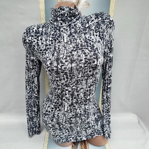 Op de afbeelding: Een nauwsluitende top met lange mouwen en een hoge, gerimpelde halslijn. De stof heeft een zwart-wit abstract patroon. De top wordt op een mannequin getoond, waarbij de getailleerde pasvorm en het ontwerp worden benadrukt.